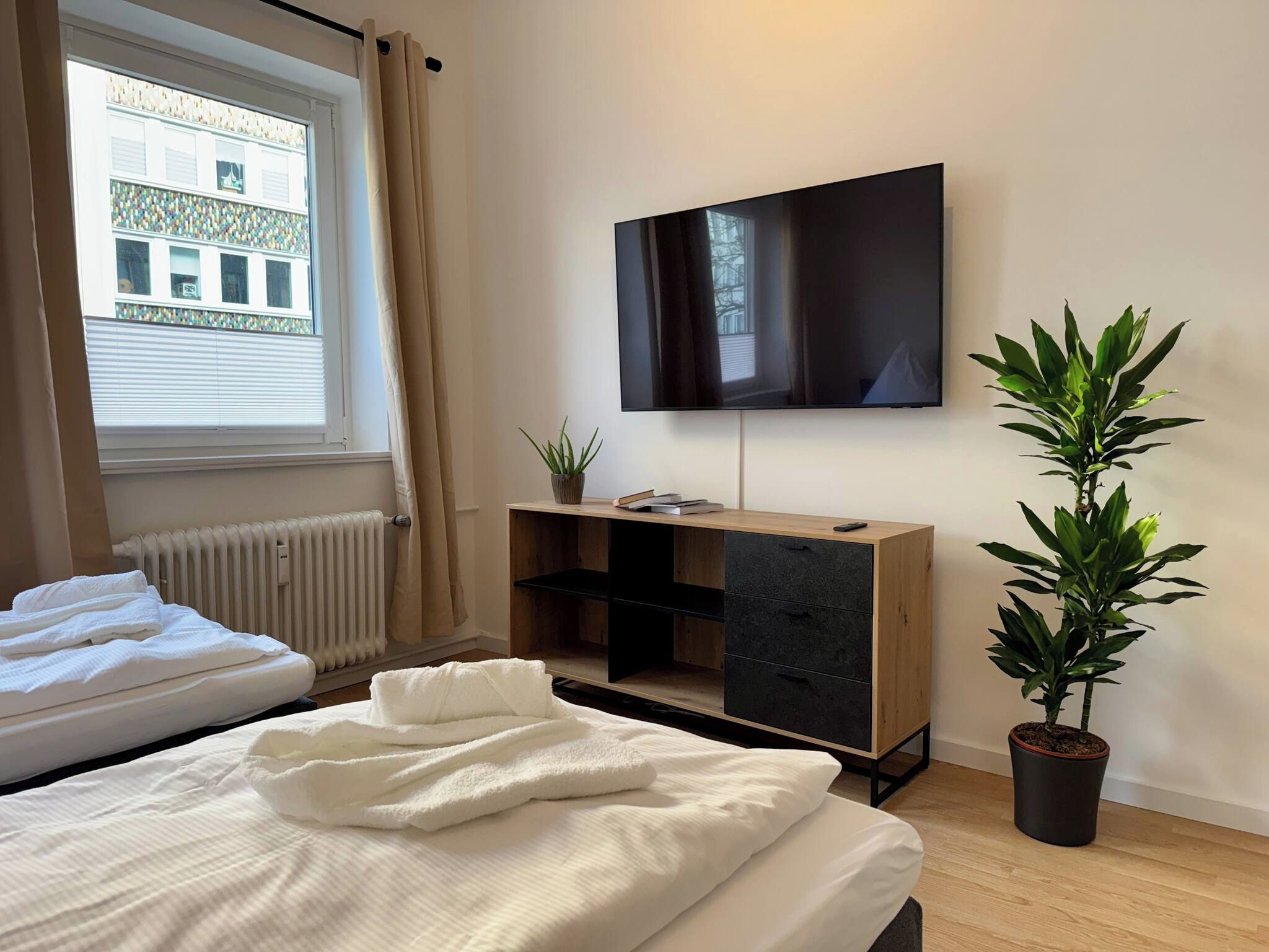 Un dormitorio con cama, televisor montado en la pared, radiador y una planta en maceta.