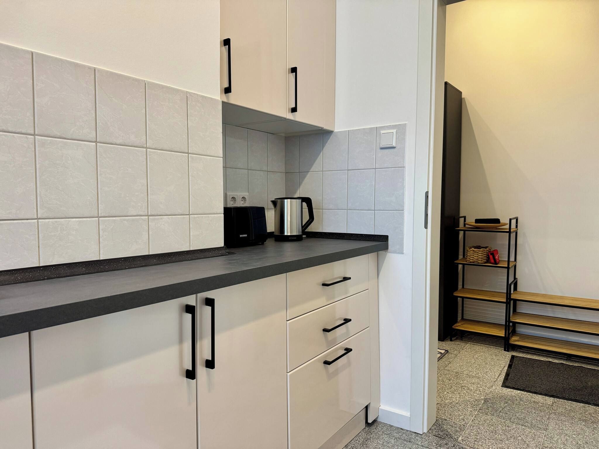 A modern kitchen with white cabinets, black handles, and a black countertop. There is a kettle on the counter and a power outlet on the wall.