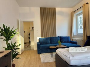 Una habitación de hotel moderna con un sofá azul, una pequeña mesa redonda, una cama blanca y una planta en maceta.