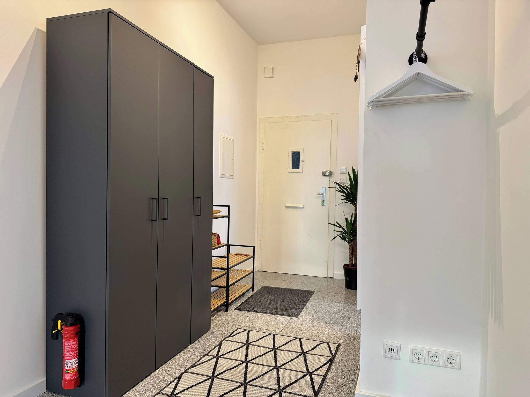 Un couloir avec une armoire, un extincteur, une étagère avec divers objets, une porte et une plante en pot.