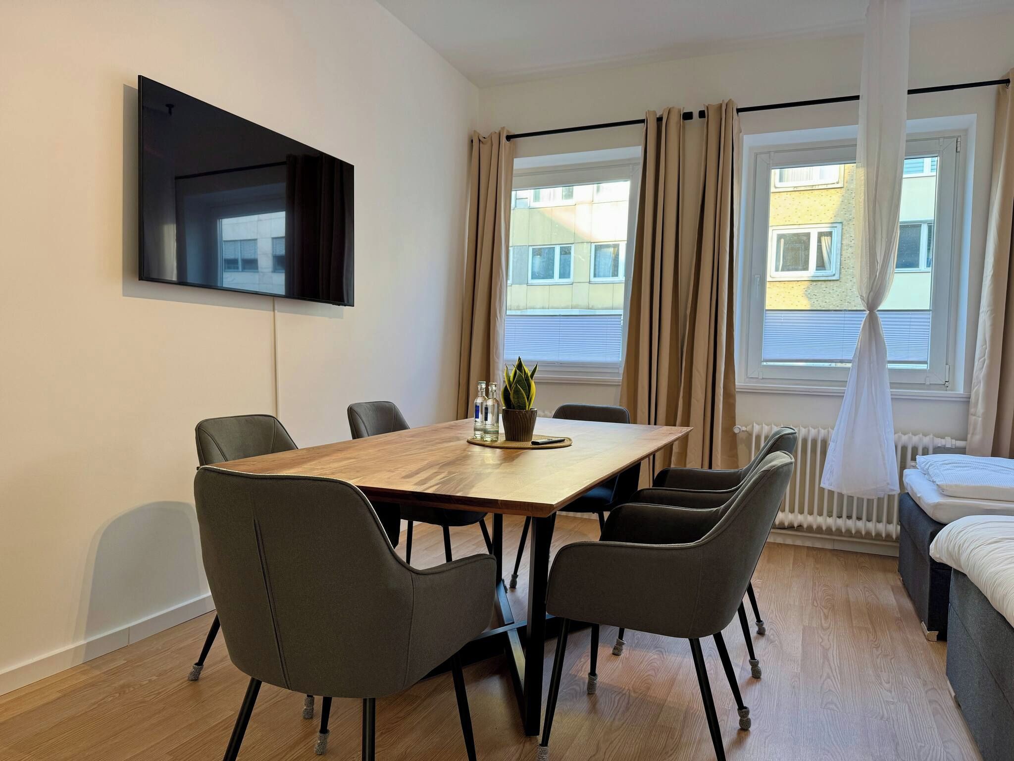 Ein modernes Hotelzimmer mit Essbereich, einem Wand-Fernseher und großen, mit Vorhängen versehenen Fenstern.