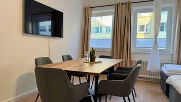 Ein modernes Hotelzimmer mit Essbereich, einem Wand-Fernseher und großen, mit Vorhängen versehenen Fenstern.