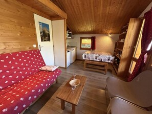 Tunnelmallinen huone, jossa on punainen sohva, puinen pöytä, jonka päällä on kaksi viinilasia, pieni keittiöalue ja ikkuna, jossa on verho.