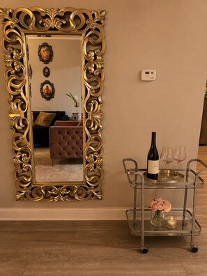 Una habitación con un espejo grande y ornamentado, un sofá, una mesita con una botella y vasos, y un carrito con un florero de flores.
