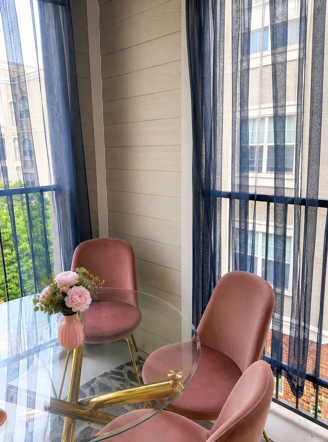 A room with a glass table, two pink chairs, a vase with flowers, and a view of a building through sheer curtains.