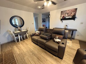 Een woonkamer met een bruine leren bank, een eettafel met stoelen, een ronde spiegel en een plafondventilator.