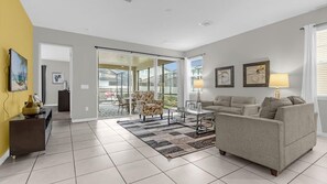 A modern living room with a sofa, armchairs, a coffee table, and a rug. There is a sliding glass door leading to a balcony with outdoor furniture. - Magical Disney Retreat with Pool, Spa & Theater (Kissimmee)