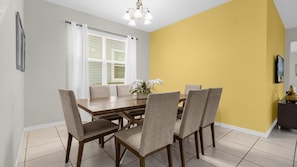 A dining area with a wooden table and six chairs, a window with blinds, and a yellow accent wall. - Magical Disney Retreat with Pool, Spa & Theater (Kissimmee)