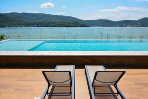 A pool with a view of a lake and mountains, two lounge chairs, and a wooden deck. - Elegante Loft Sunset Lagoa - LDI006 (Florianópolis)