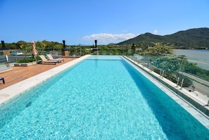 A rectangular swimming pool with clear blue water, surrounded by a wooden deck and lounge chairs. - Elegante Loft Sunset Lagoa - LDI006 (Florianópolis)