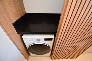 A compact laundry area with a washing machine and a black countertop. - Elegante Loft Sunset Lagoa - LDI006 (Florianópolis)