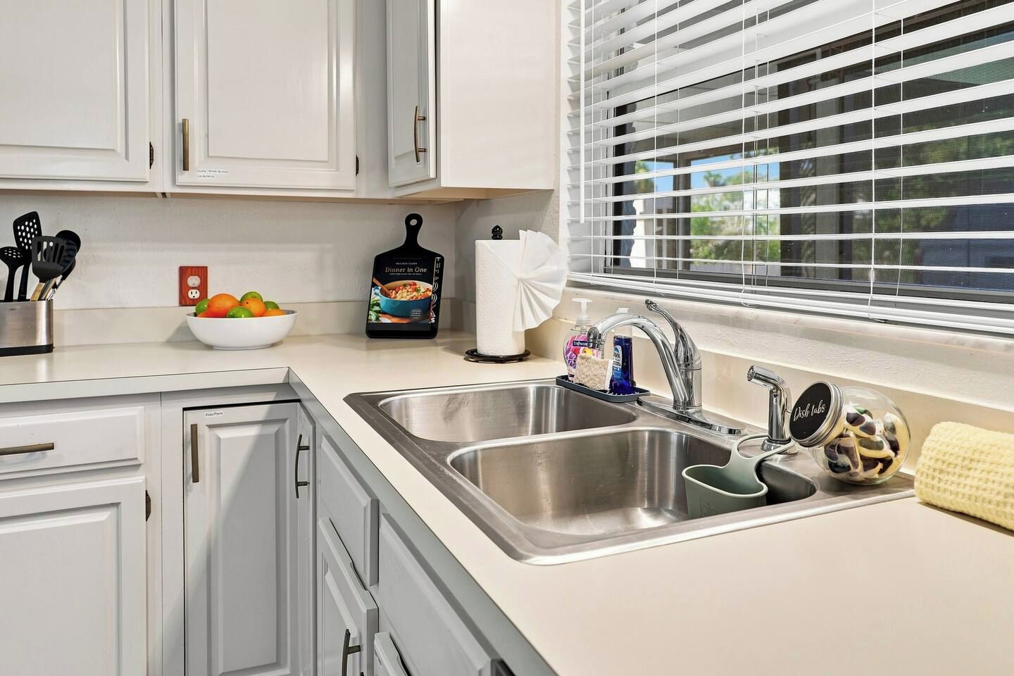 A kitchen with white cabinets, a stainless steel sink, a bowl of fruit, a bottle of dish soap, and a window with blinds.