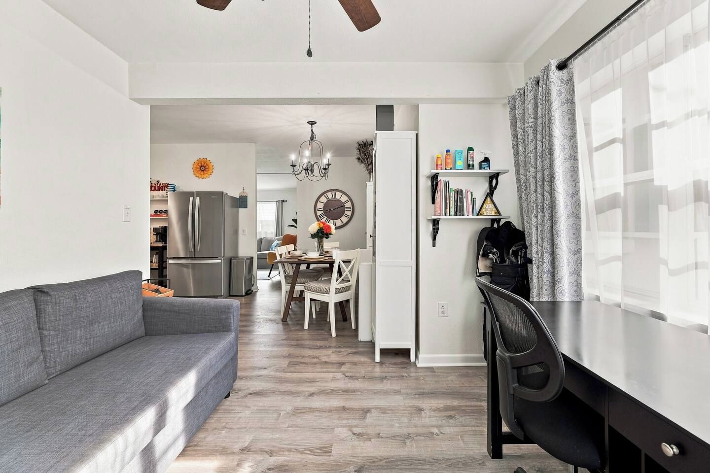 A modern living room with a gray sofa, a dining area with a chandelier, and a kitchen with a refrigerator.