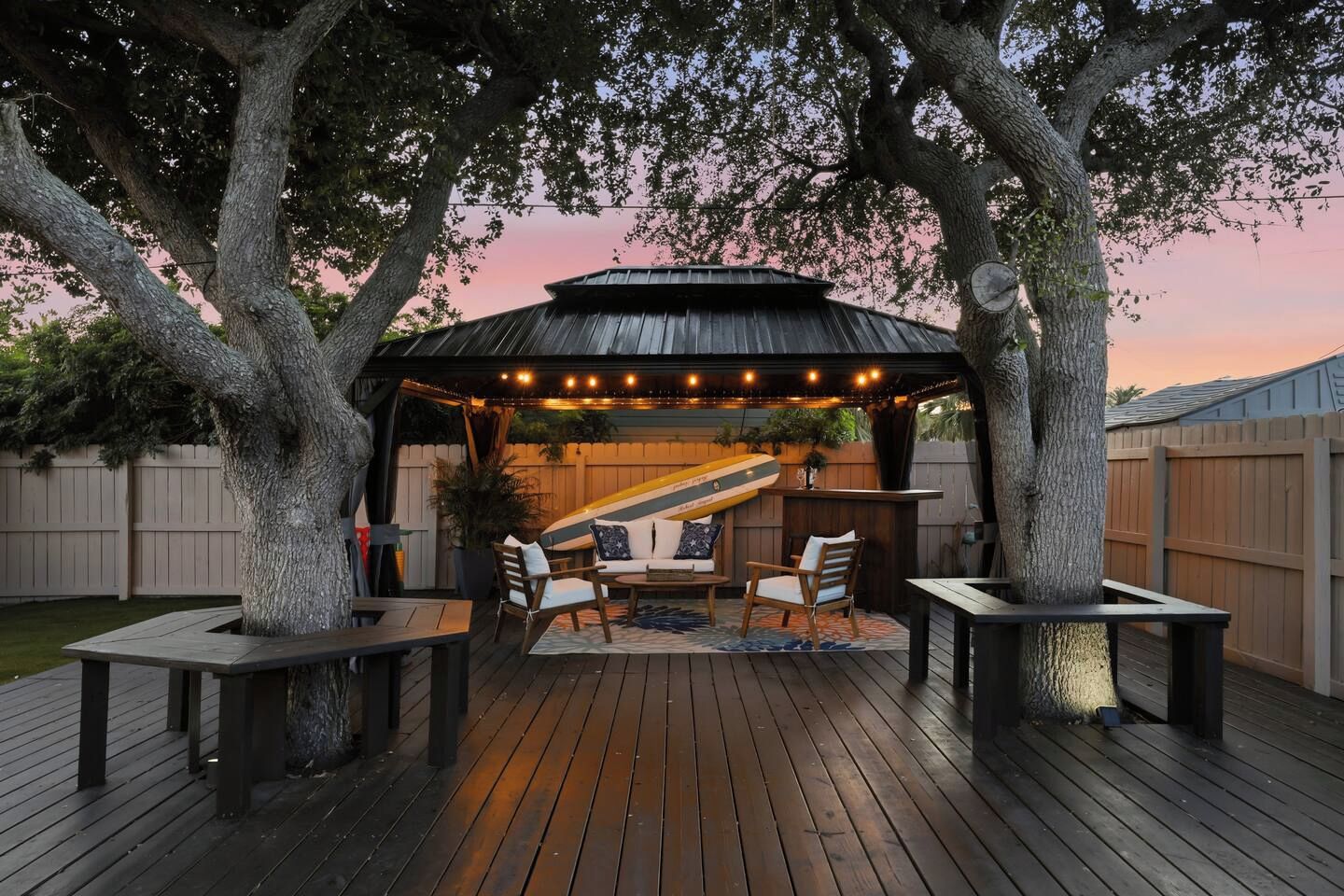A covered outdoor seating area with a table and chairs, surrounded by trees and a wooden deck.
