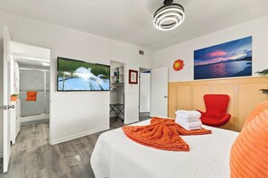 Ett hotellrum med en säng, en röd stol, en tv och en tavla som föreställer en solnedgång.