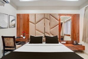 A modern hotel room with a large bed, a wooden headboard, a desk with a chair, a vase of flowers, and a window with curtains.