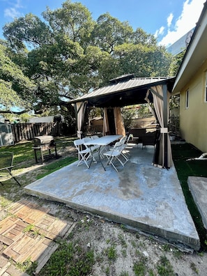 Een overdekt terras met een tafel, stoelen, een barbecue en een omheind gazon.