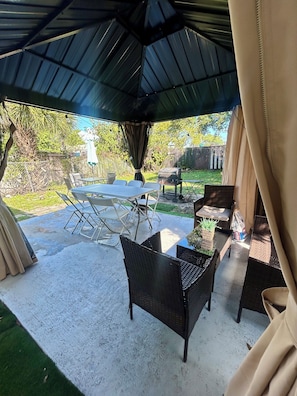 Un patio techado al aire libre con mesa y sillas, una parrilla y vista al jardín trasero.