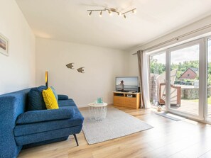 A modern living room with a blue sofa, a television on a wooden stand, and a large glass door leading to an outdoor area. - 1 "coastal Child" (St. Peter-Ording)