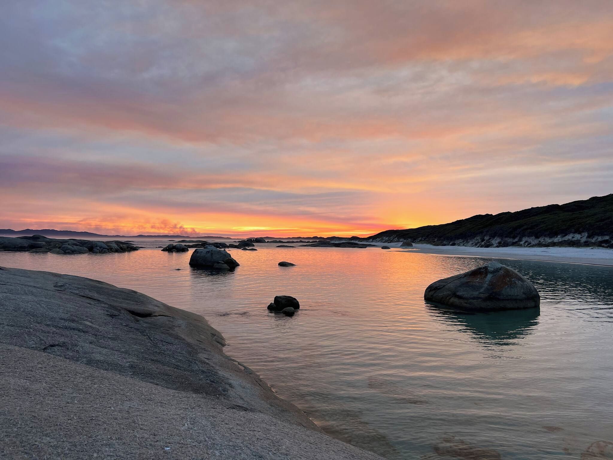 A coastal landscape with calm water, large rocks, and a distant shoreline during sunset.