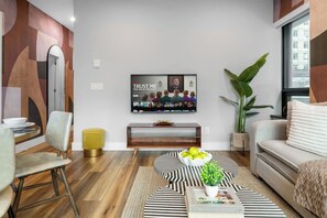 Una sala moderna con una televisión de pantalla plana montada en la pared, una mesa de centro de madera con un tazón de manzanas, un sofá y una planta grande.