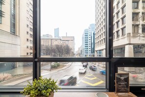 Blick aus einem Fenster mit einem Blumentopf auf der Fensterbank, einem Schild, das kostenloses WLAN anzeigt, und belebter Straße mit Autos und Gebäuden im Hintergrund.