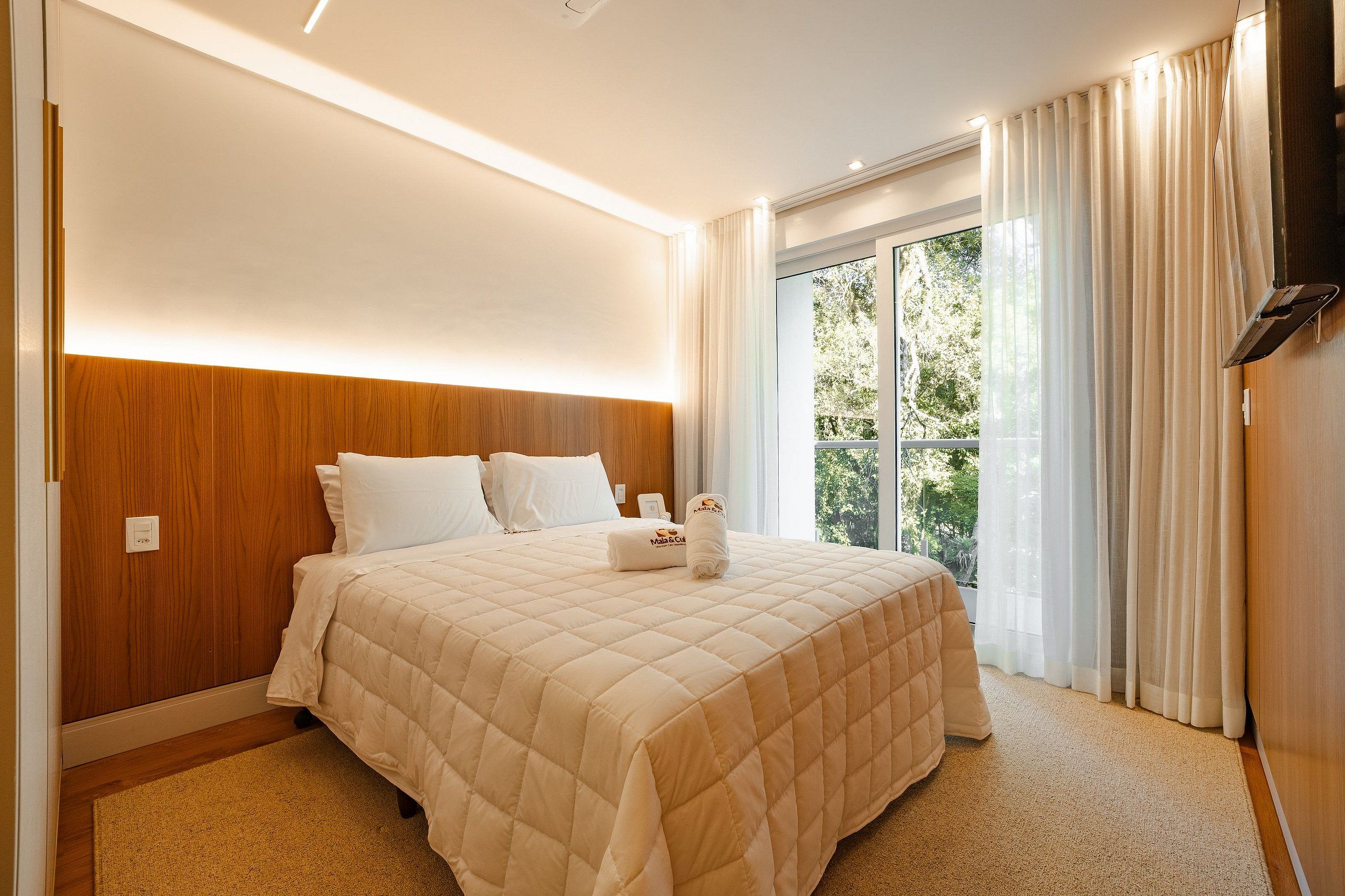 A hotel room with a large bed, a wooden headboard, a flat-screen TV, and a window with curtains.