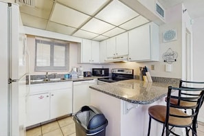 A kitchen with white cabinets, a granite countertop, a stove, a microwave, and a trash can. - Sunbird 1107e (Panama City Beach)