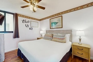 A hotel room with a bed, two bedside tables with lamps, a ceiling fan, a window with curtains, a framed quote on the wall, and a framed picture above the bed. - Sunbird 1107e (Panama City Beach)