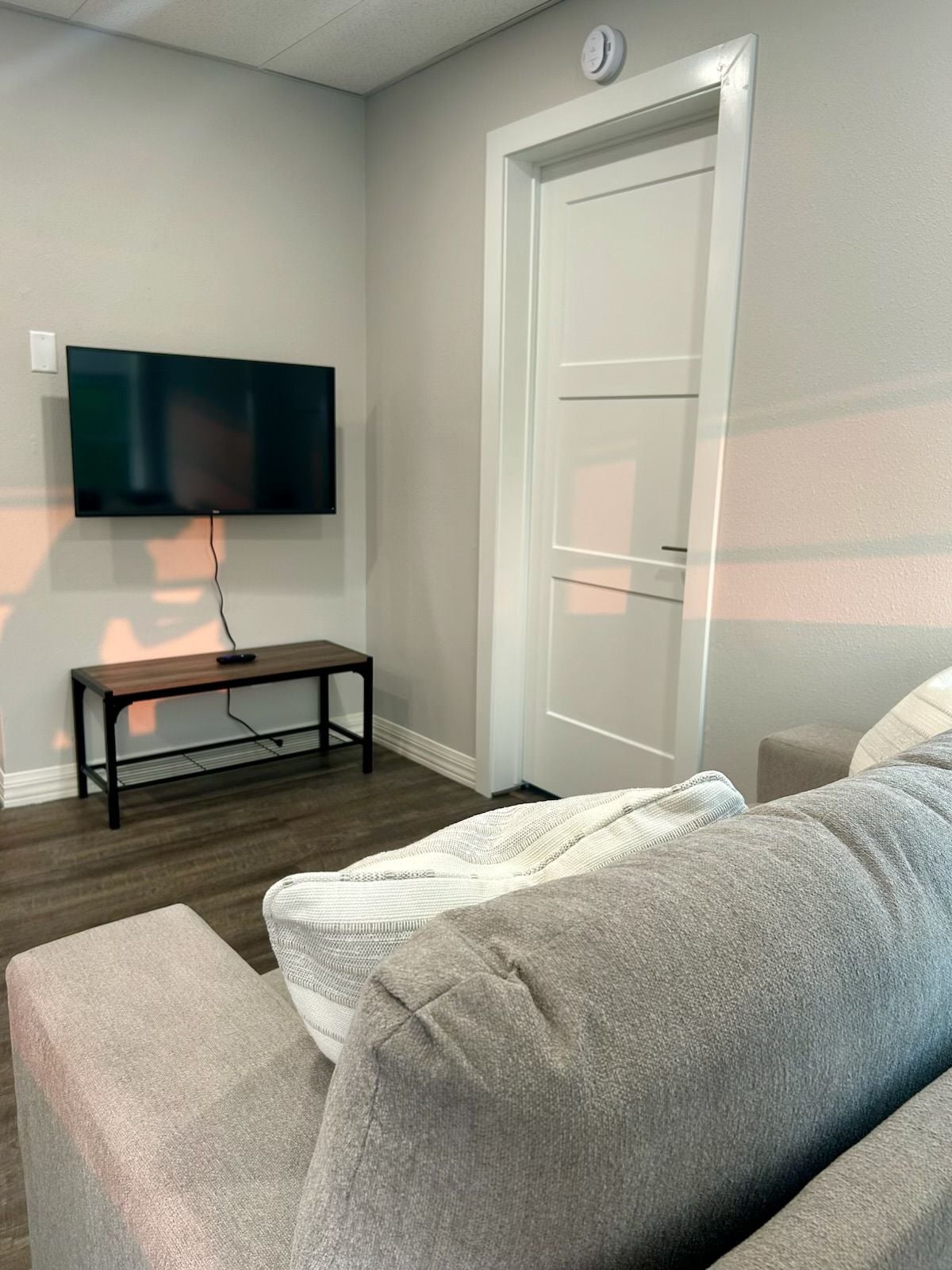 A living room with a flat-screen TV mounted on the wall, a wooden coffee table, a grey sofa, and a white door.