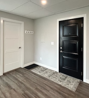 A hallway with a white door, a black door, a small rug, and a light switch on the wall.