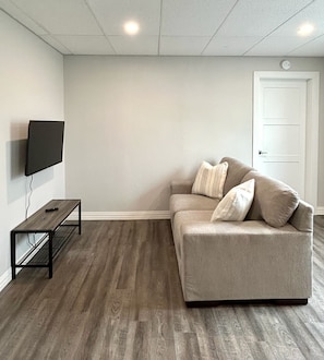 A room with a grey sofa, a flat-screen TV mounted on the wall, a small black table, and a white door.