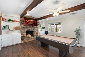 Una mesa de billar en una habitación con chimenea de piedra, vigas de madera y ventilador de techo.