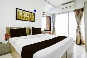 Ein Hotelzimmer mit einem großen Bett, einem Nachttisch, einem kleinen Schreibtisch mit Stuhl, einem Fenster mit Vorhängen und einer Wand-Klimaanlage.