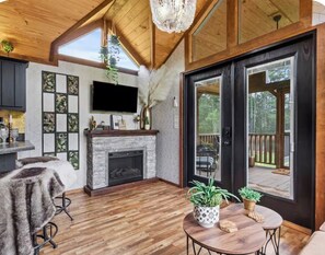 A spacious living area with a fireplace, wooden flooring, and a large window overlooking a deck. - Where peace meets the pines!! Pond fishing, fire pit & egg chairs! (Boaz)