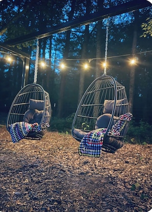 Two hanging chairs suspended from a wooden structure, with a string of lights above. - Where peace meets the pines!! Pond fishing, fire pit & egg chairs! (Boaz)