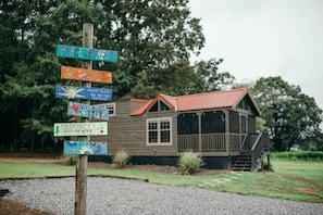 A wooden cabin with a red roof and a porch, surrounded by a gravel area and trees.