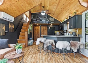 A rustic cabin interior with a wooden ceiling, a kitchen area, and a seating area with stools.