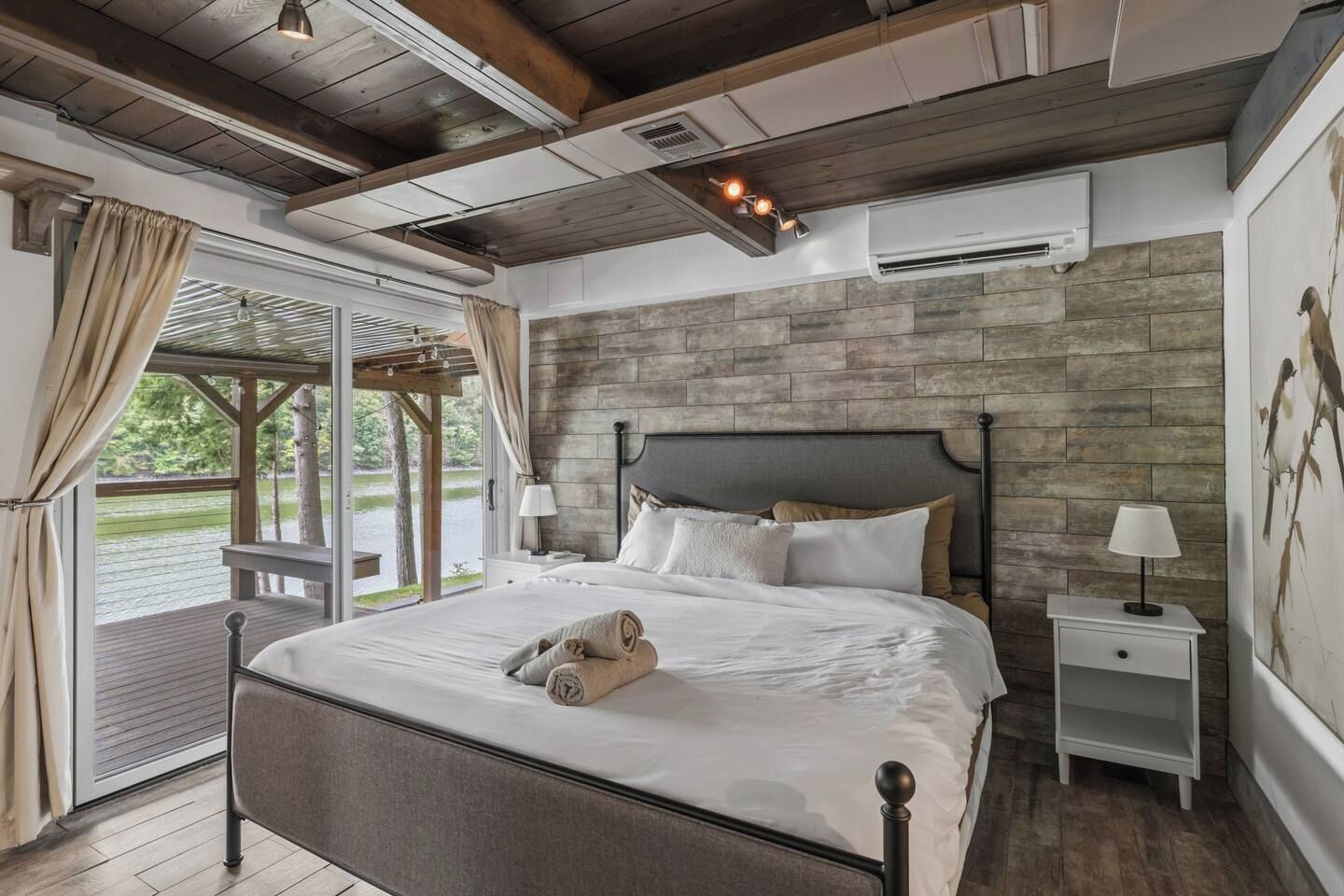A bedroom with a large bed, a wooden ceiling, a stone wall, a window with curtains, and a nightstand with a lamp.