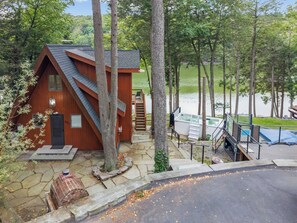 A cabin with a stone pathway leading to a wooden deck, surrounded by trees and a body of water. - Lakefront Escape | Hot Tub, Theater & Game Room (Monticello)