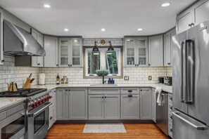 A modern kitchen with grey cabinets, stainless steel appliances, a window with a view of trees, and a wooden floor. - Lakefront Escape | Hot Tub, Theater & Game Room (Monticello)