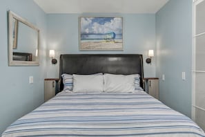 Ein Schlafzimmer mit einem Bett, einem Kopfteil, zwei Wandlampen, einem Bilderrahmen mit einer Strandansicht und einem Spiegel.
