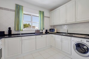 A modern kitchen with white cabinets, a built-in washing machine, a microwave, and a window with curtains. - Casa da Galé | Pool & Terrace (Albufeira)
