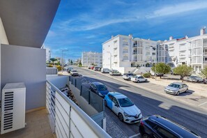 Rua residencial com edifícios brancos, carros estacionados e céu claro.