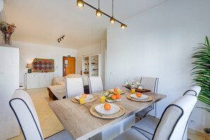 Ein Essbereich mit einem Holztisch, gedeckt für vier Personen, umgeben von Stühlen. Rechts ist eine Pflanze und links ein weißes Sideboard mit gemustertem Tuch zu sehen.