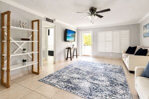 Ein Wohnzimmer mit einem weißen Sofa, einem blauen Teppich mit Muster, einem Deckenventilator und einem an der Wand befestigten Fernseher.