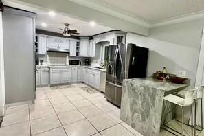 A kitchen with white cabinets, a refrigerator, a microwave, and a bar area with stools. - CL6900-3D-Private Heated pool home in a quiet area (Fort Lauderdale)