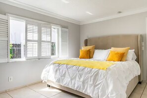 Ein Schlafzimmer mit einem großen Bett, weißer Bettwäsche und Blick durch ein Fenster mit Jalousien.