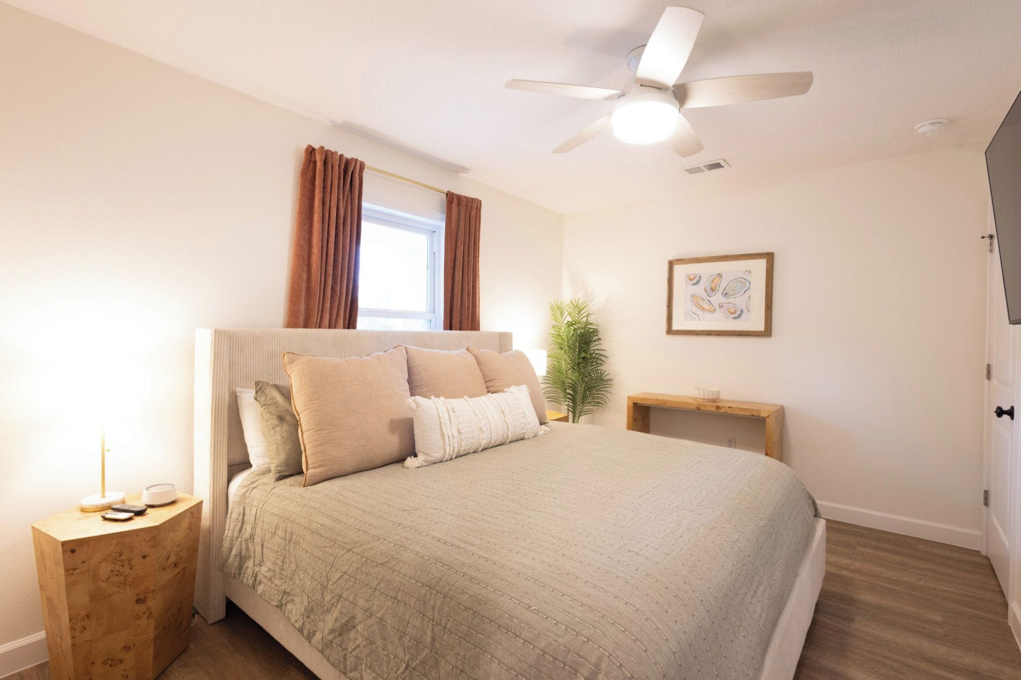 A bedroom with a bed, a ceiling fan, a window with curtains, a bedside table, a nightstand, a plant, and a framed picture on the wall.