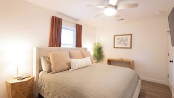 A bedroom with a bed, a ceiling fan, a window with curtains, a bedside table, a nightstand, a plant, and a framed picture on the wall.